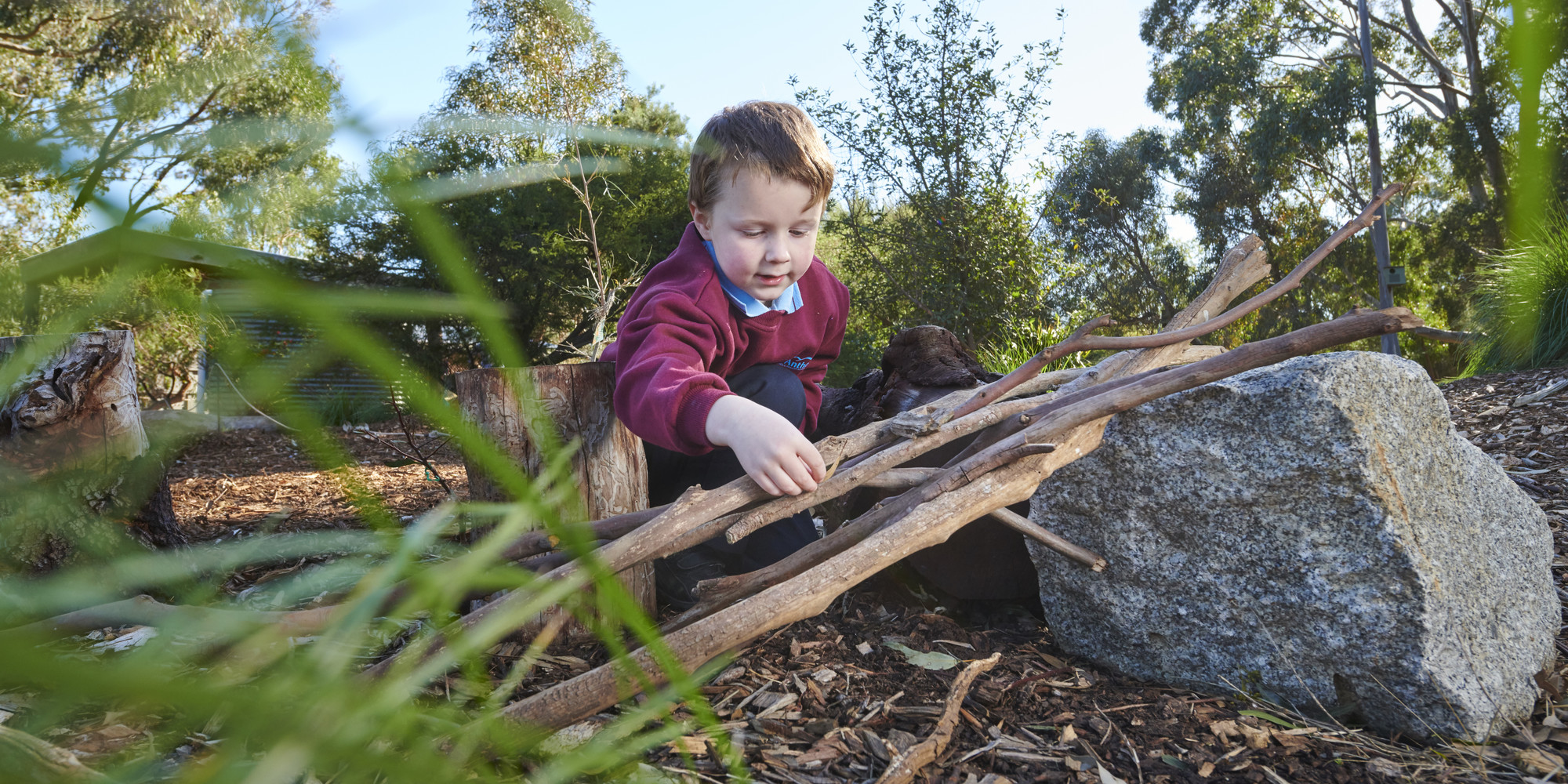 A young school student is crouched on the ground stacking some sticks.