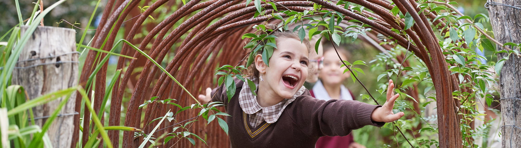 Girl in brown school uniform smiles as she walks through an archway and two other students can be seen behind her.