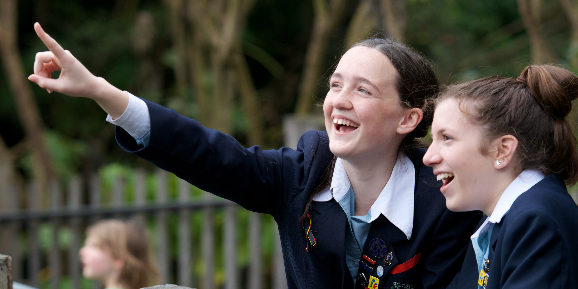 Two teen girls dressed in school uniform look excitedly at something out of frame, one pointing left.